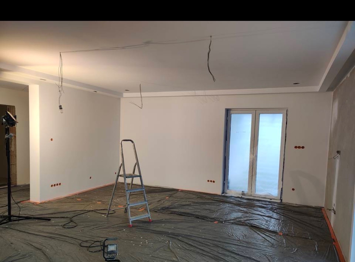 Przygotowane do malowania pomieszczenie z zabezpieczoną folią podłogą, aluminiową drabiną, lampą budowlaną i wystającymi przewodami elektrycznymi ze ściany i sufitu.