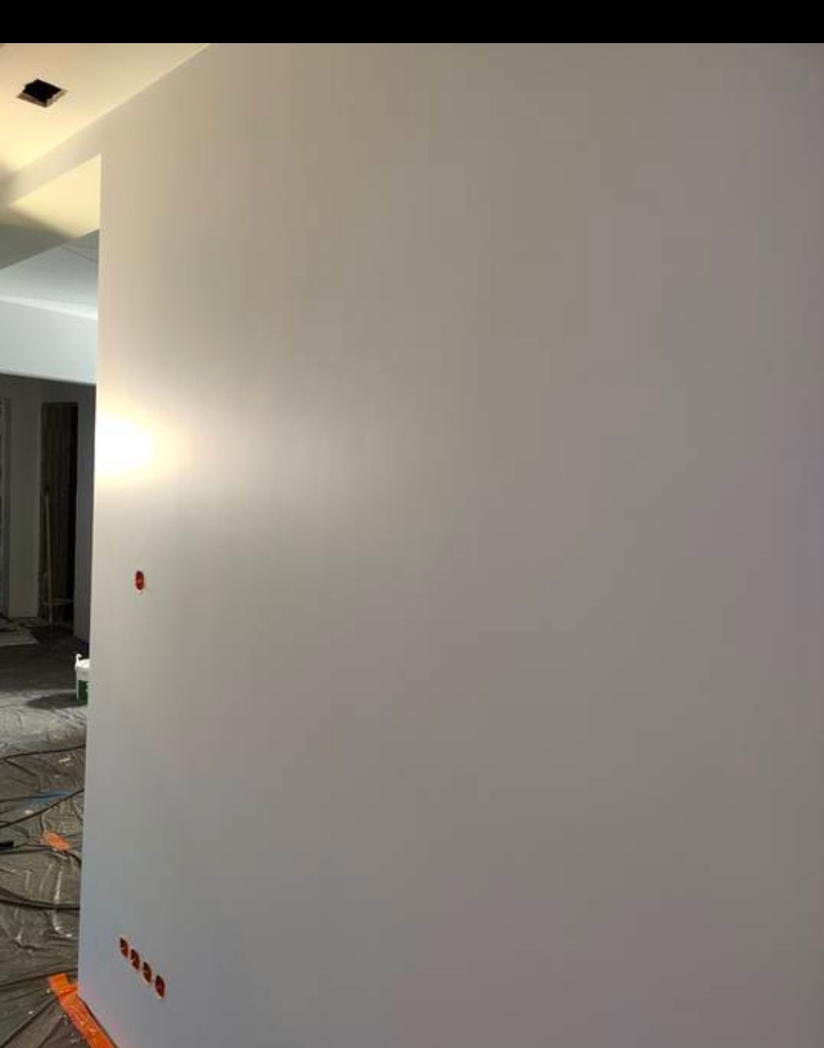 Świeżo wyszpachlowana ściana pomalowana na jasny odcień szarości, widoczne gniazdka elektryczne zabezpieczone czerwonymi zaślepkami, podłoga zabezpieczona folią malarską.