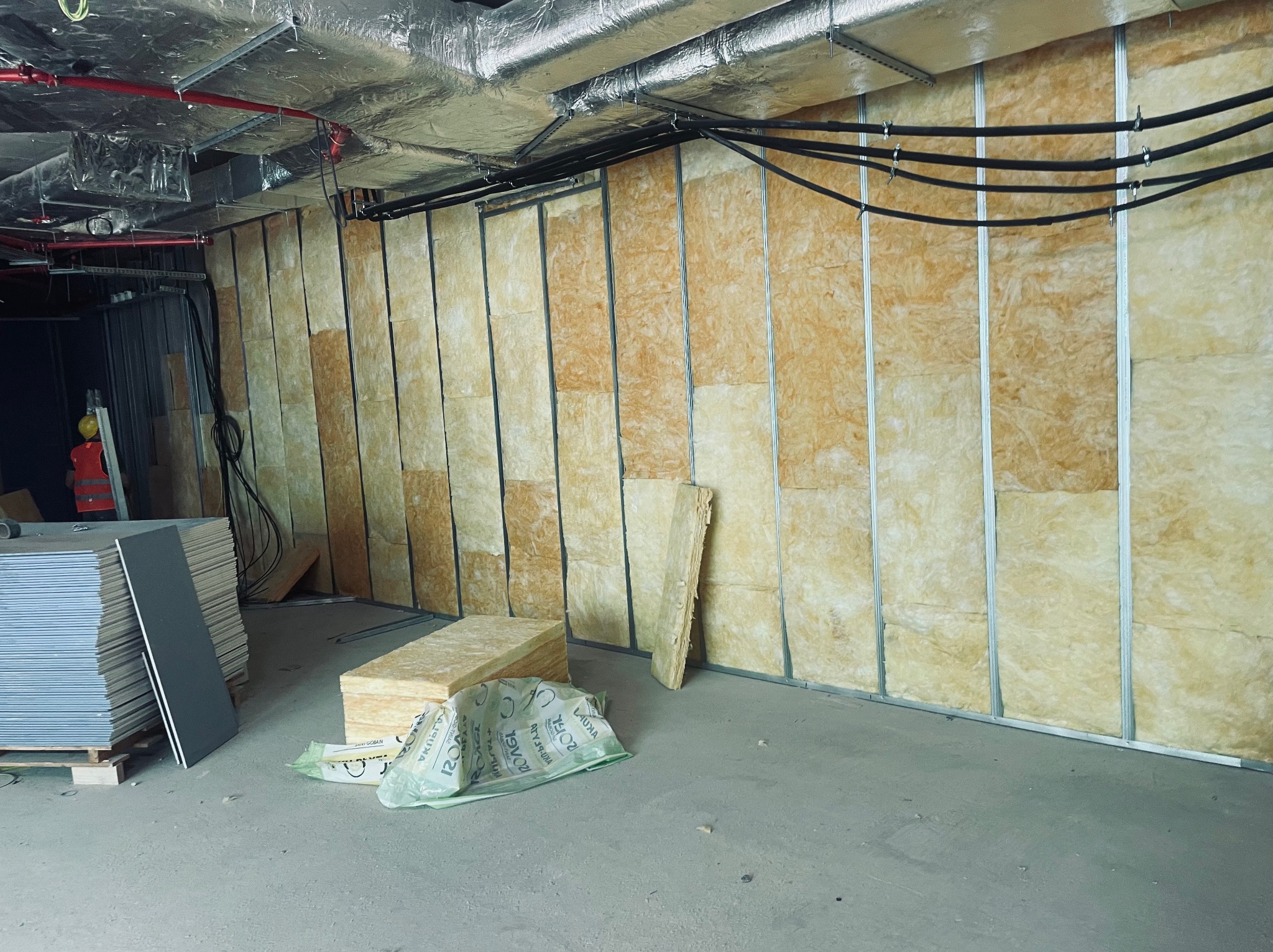 Montaż izolacji z wełny mineralnej w metalowym stelażu ścian działowych, widoczne płyty kartonowo-gipsowe na palecie i materiały izolacyjne w folii, instalacje pod sufitem.