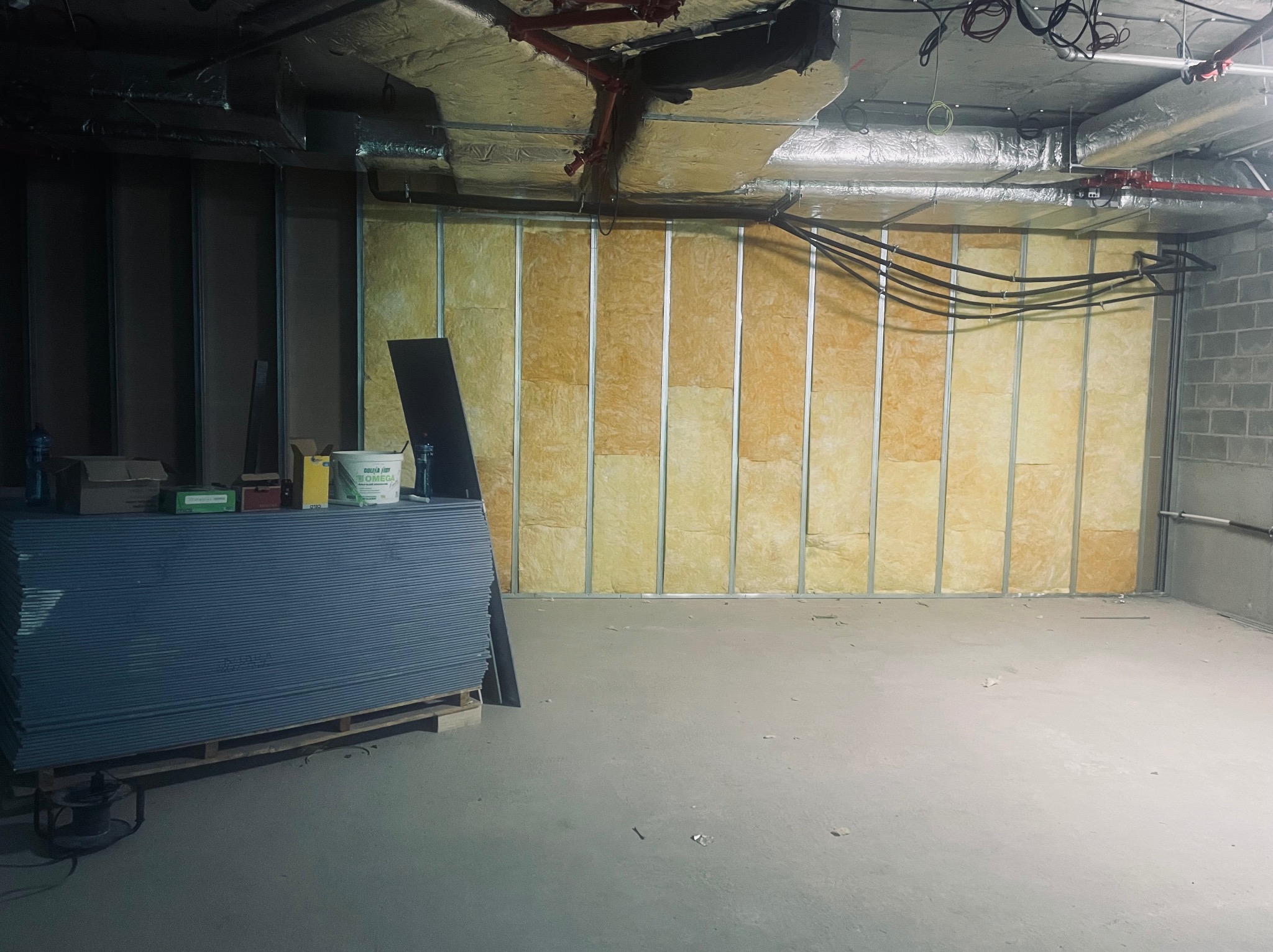 Stos płyt kartonowo-gipsowych na palecie w surowym wnętrzu z metalowym szkieletem ścian wypełnionym izolacją z wełny mineralnej, widoczne instalacje pod sufitem.