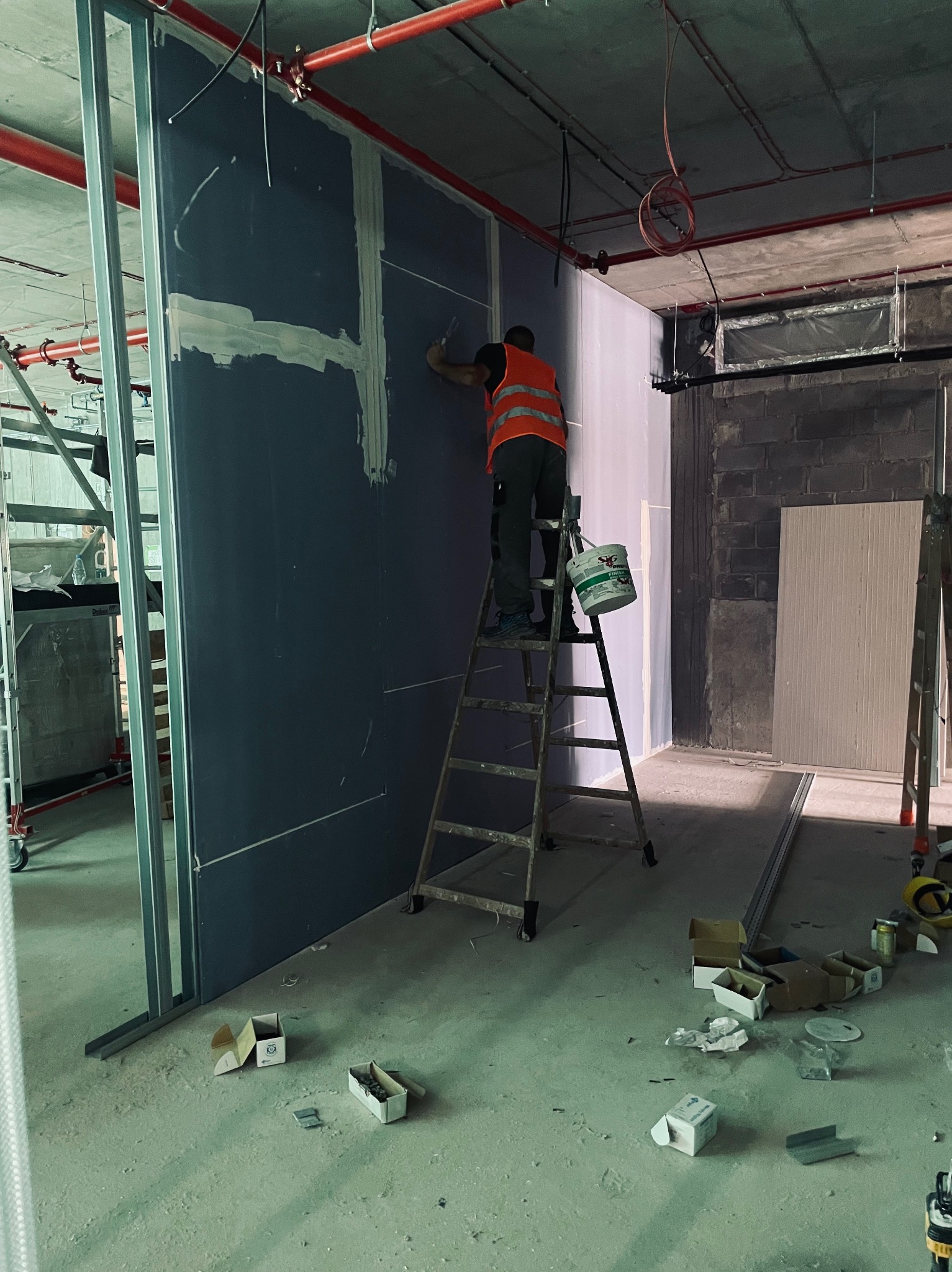 Pracownik w pomarańczowej kamizelce nakłada gładź na szarą płytę karton-gips stojąc na metalowej drabinie w surowym wnętrzu, widoczna puszka z gładzią, metalowe profile i akcesoria na podłodze.
