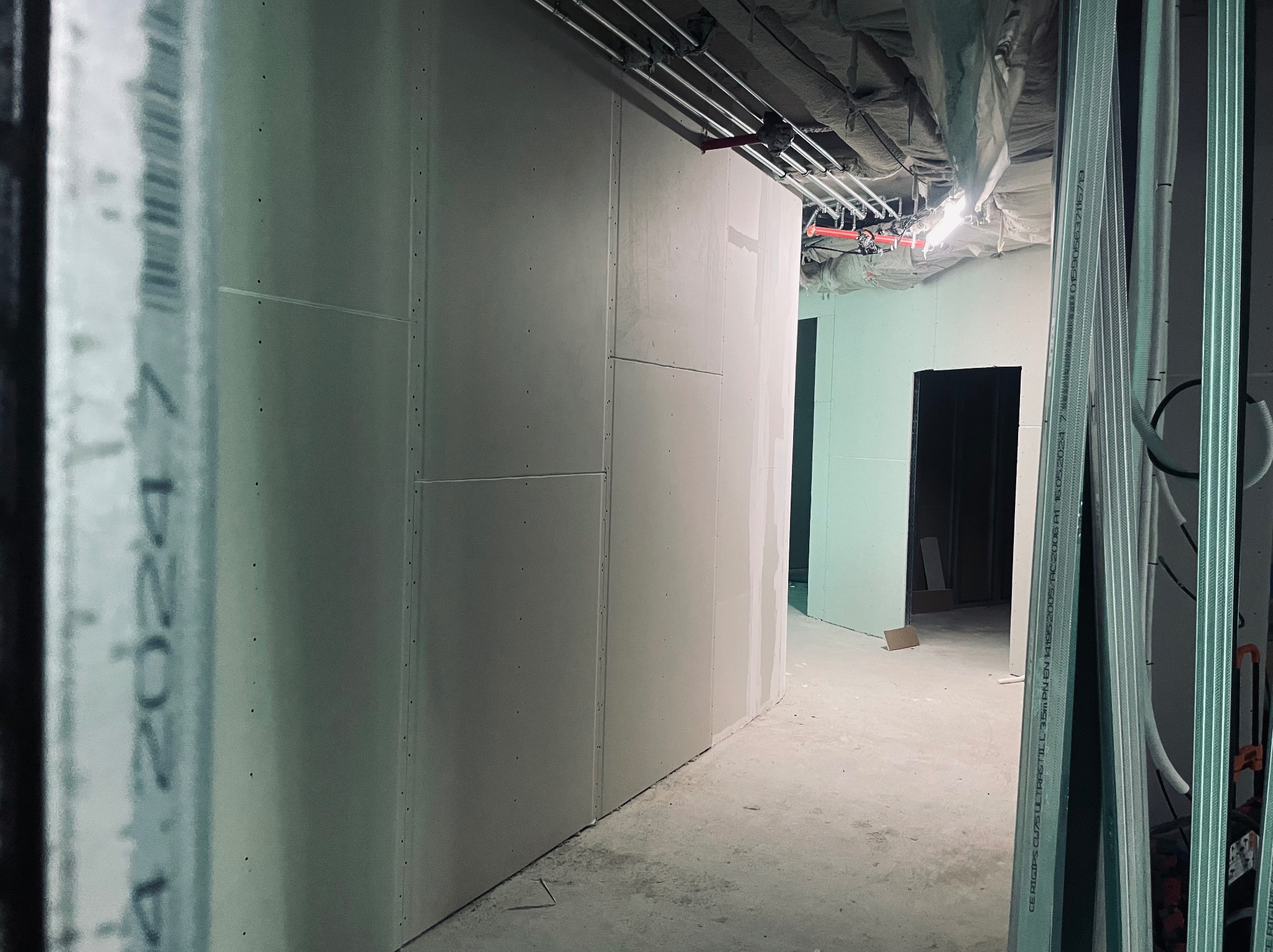 Wnętrze w trakcie prac wykończeniowych: zamontowane płyty gipsowo-kartonowe na ścianach, widoczne metalowe profile konstrukcyjne, instalacje pod sufitem i surowy beton na podłodze, korytarz...