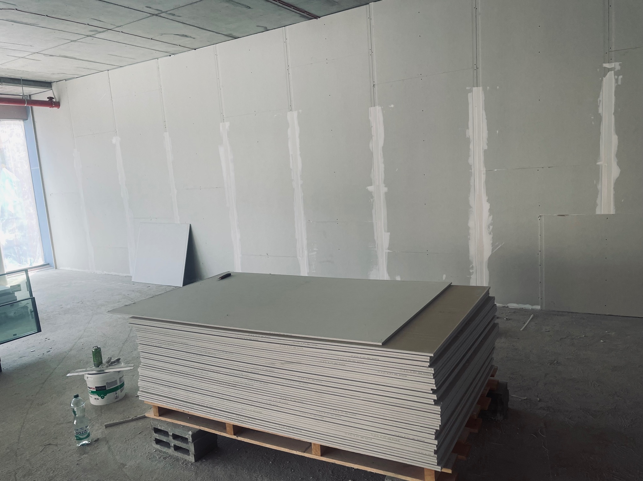 Stos płyt gipsowo-kartonowych na palecie w surowym wnętrzu, widoczne łączenia płyt na ścianie, wiadro z zaprawą i narzędzia obok.
