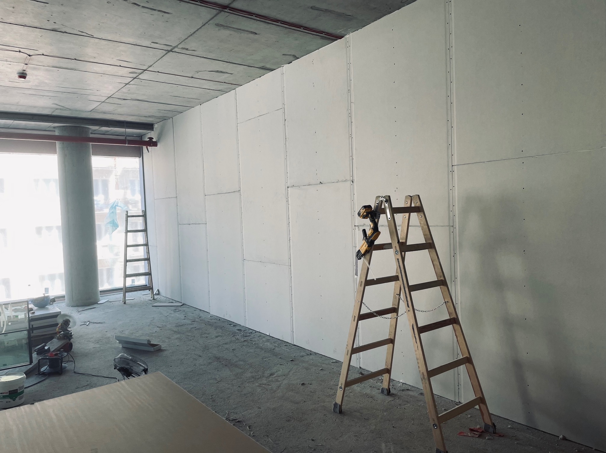Montaż ścian z płyt gipsowo-kartonowych w surowym wnętrzu biurowym, widoczna drewniana drabina z wkrętarką, betonowy sufit i okno z widokiem na miasto.
