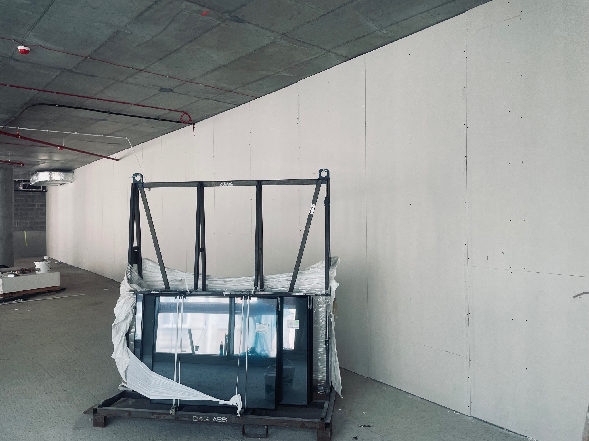 Nowe okna w folii ochronnej, ustawione na metalowym stojaku przed ścianą z płyt kartonowo-gipsowych w surowym wnętrzu budynku w trakcie wykończenia.