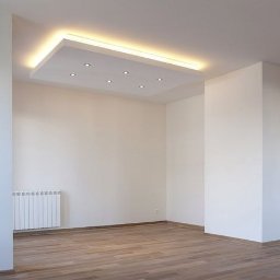 Mich-bud - Puste, jasne wnętrze z drewnianą podłogą, białymi ścianami i sufitem podwieszanym z oświetleniem punktowym i listwą LED, widoczny grzejnik pod oknem.