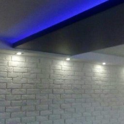 Mich-bud - Ściana z białej cegły dekoracyjnej z oświetleniem punktowym i podświetlanym na niebiesko sufitem w pomieszczeniu w Rawie Mazowieckiej.