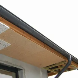 Fragment elewacji w trakcie ocieplania: widoczne płyty izolacyjne, drewniana konstrukcja podbitki, rynna i fragment okna. Dwa białe przewody elektryczne zwisają spod podbitki.