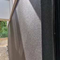 Szara elewacja budynku z widocznym efektem cieniowania światła słonecznego, fragment okna po prawej stronie i widok na ogród w tle.