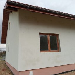 Mich-bud - Świeżo otynkowana elewacja domu jednorodzinnego w Rawie Mazowieckiej, widoczne brązowe okna i dach, elewacja w kolorze jasnego beżu, na dole cokół w kolorze brązowym, na tle pochmurne niebo.