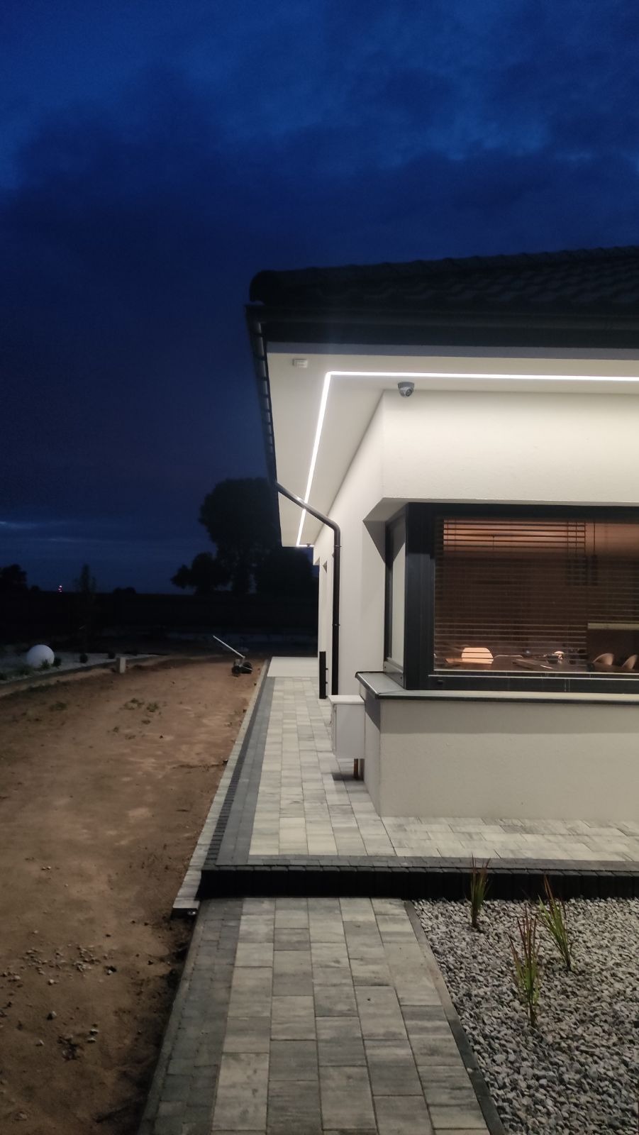 Nowoczesny dom z białą elewacją oświetloną ledowym paskiem pod okapem, widoczny system rynnowy, okno z żaluzjami i ścieżka z kostki brukowej, tło stanowi wieczorne niebo.
