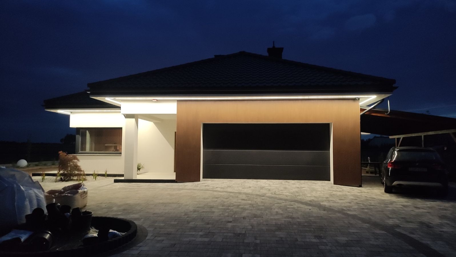 Nowoczesny dom jednorodzinny z oświetloną elewacją i automatyczną bramą garażową w wieczornym oświetleniu, widoczne worki z materiałami budowlanymi na podjeździe.