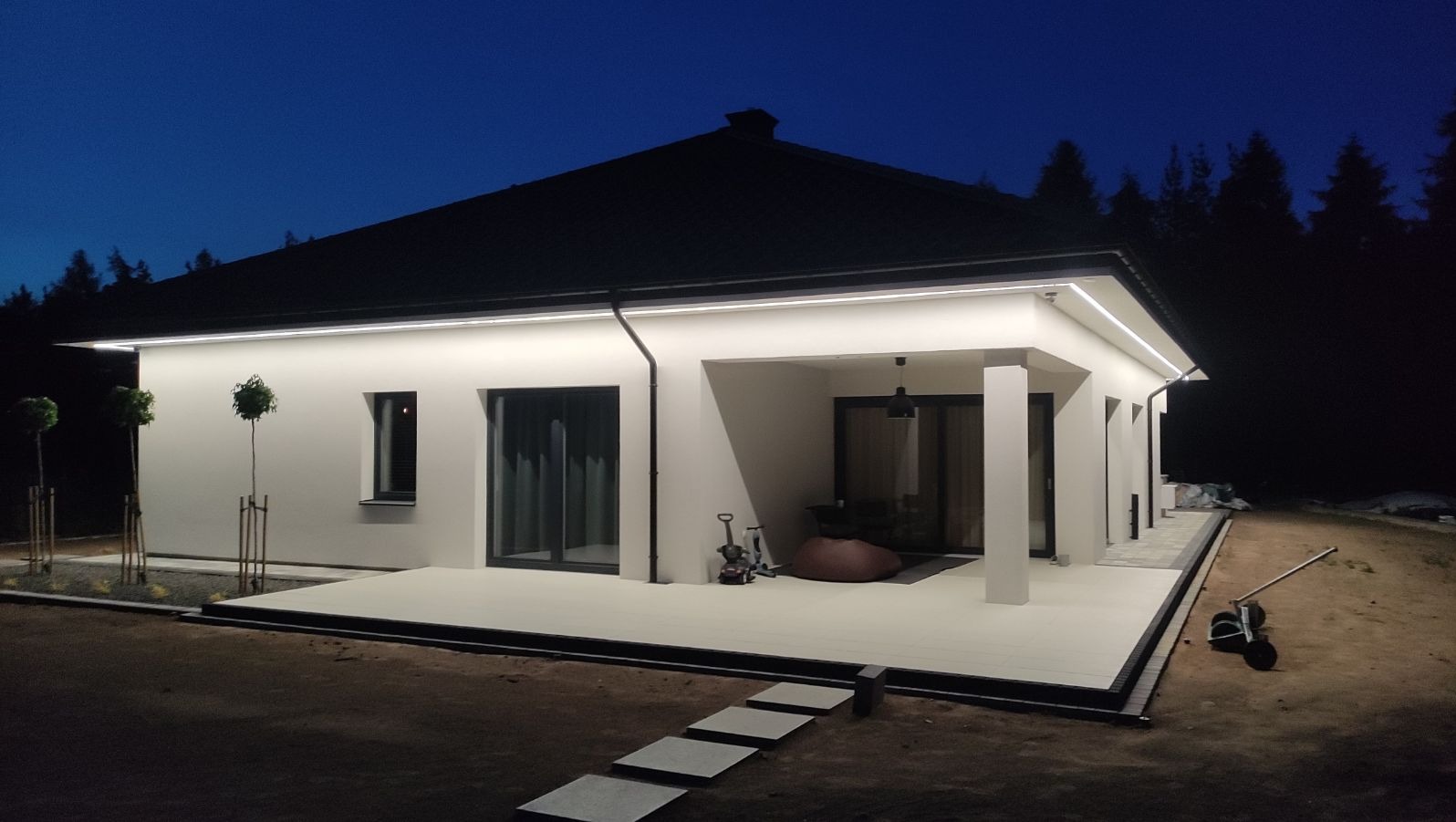 Nowoczesny dom jednorodzinny z białymi ścianami, podświetloną elewacją i tarasem, sfotografowany o zmierzchu z widocznymi elementami ogrodu i lasem w tle.