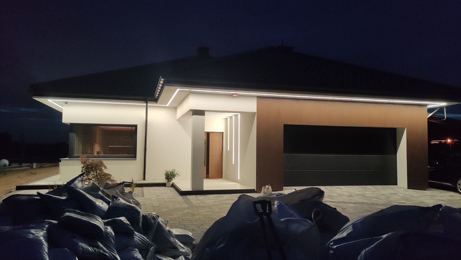 Nowoczesny dom jednorodzinny z oświetleniem elewacyjnym LED, widoczny wieczorem, z workami materiałów budowlanych na pierwszym planie.