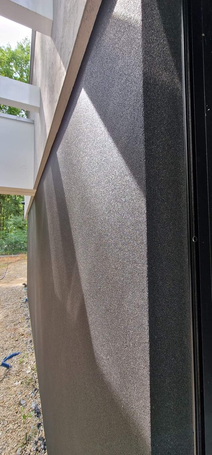 Szara elewacja budynku z widocznym efektem cieniowania światła słonecznego, fragment okna po prawej stronie i widok na ogród w tle.