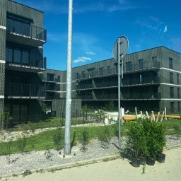 BRAVO IHOR ROZHKO - Nowoczesny budynek wielorodzinny z ciemnej cegły z balkonami, widoczny z poziomu chodnika, z elementami zagospodarowania terenu i odbiciem samochodu w szybie.