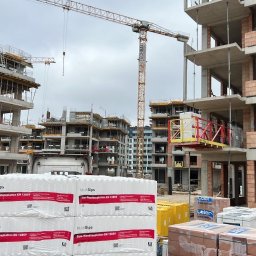 BRAVO IHOR ROZHKO - Budowa wielopiętrowego budynku z cegły i betonu, widoczne rusztowania, żuraw budowlany, palety z materiałami budowlanymi, w tym białe paczki MultiGips Gips-Wandbauplatten EN 12859.