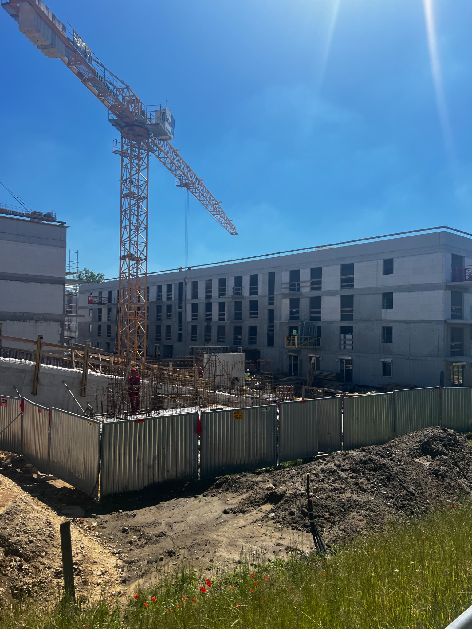 Wysoki żuraw budowlany na tle surowego, wielopiętrowego budynku w trakcie budowy. Widoczne rusztowania i robotnicy w oddali. Teren budowy zabezpieczony metalowym ogrodzeniem, w tle hałda ziemi...