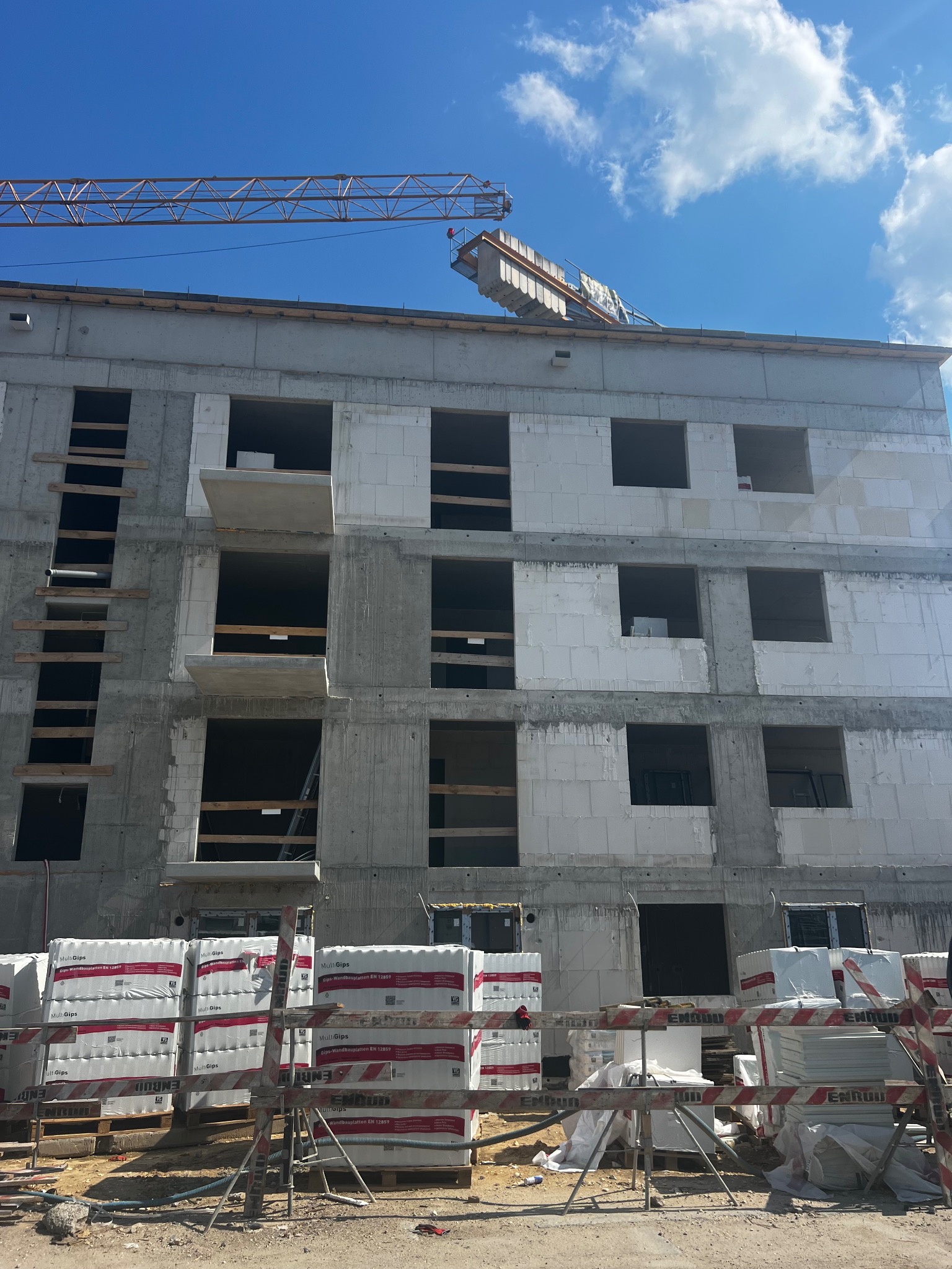 Wznoszony budynek wielorodzinny z betonu komórkowego, widoczne palety z materiałem budowlanym MultiGips na pierwszym planie oraz żuraw budowlany w tle na tle błękitnego nieba.