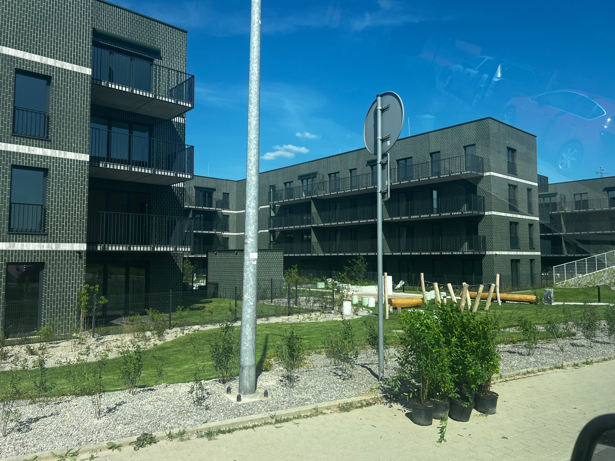 Nowoczesny budynek wielorodzinny z ciemnej cegły z balkonami, widoczny z poziomu chodnika, z elementami zagospodarowania terenu i odbiciem samochodu w szybie.