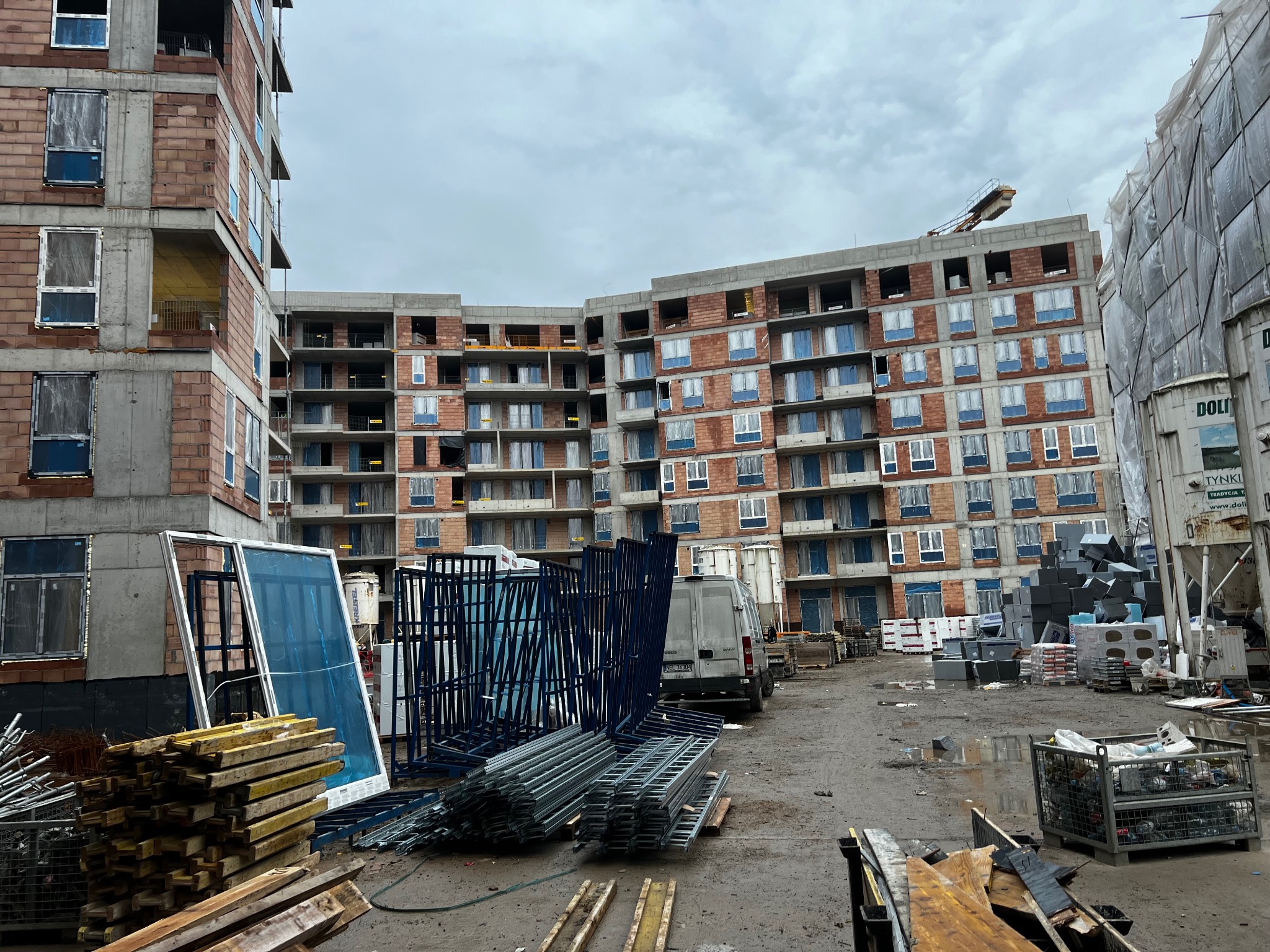 Budowa wielopiętrowego budynku z czerwonej cegły, widoczne rusztowania, okna zabezpieczone folią, stosy drewna i materiałów budowlanych na placu budowy w pochmurny dzień.