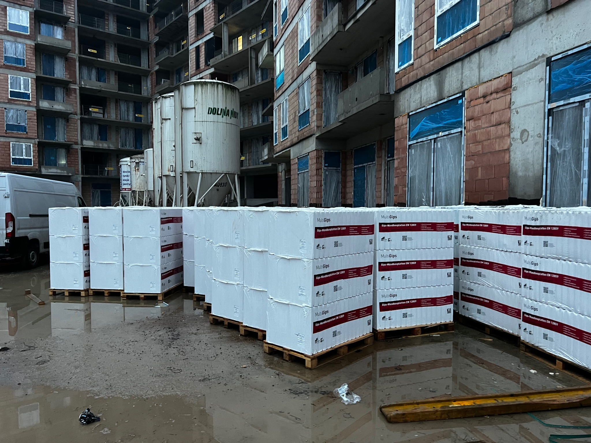 Palety z płytami gipsowymi MultiGips na mokrym placu budowy, w tle surowy budynek wielorodzinny z cegły i zbiorniki Dolina Nidy.