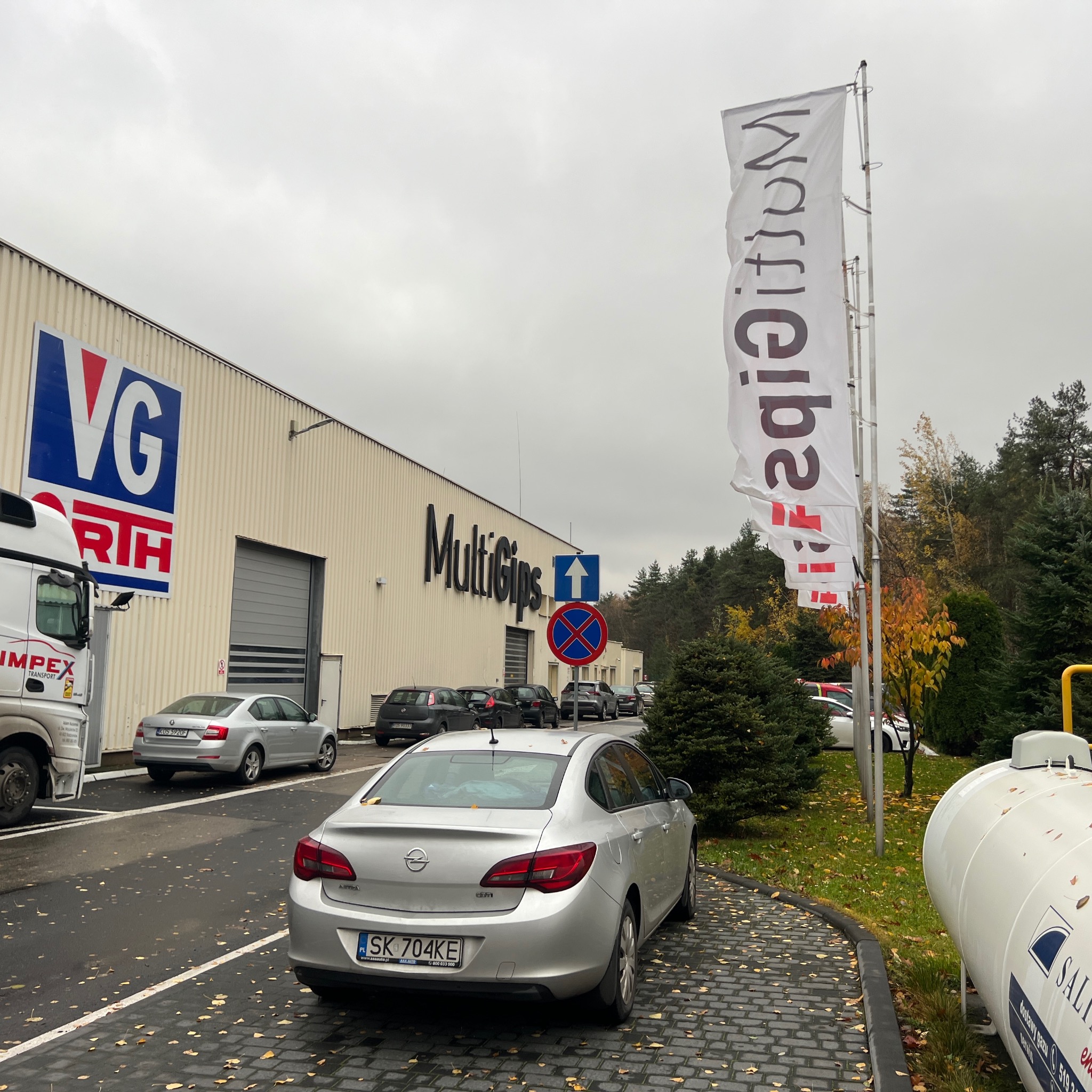 Budynek firmy VG-Orth i MultiGips z zaparkowanymi samochodami, flagi reklamowe na wietrze, jesienne liście na kostce brukowej.