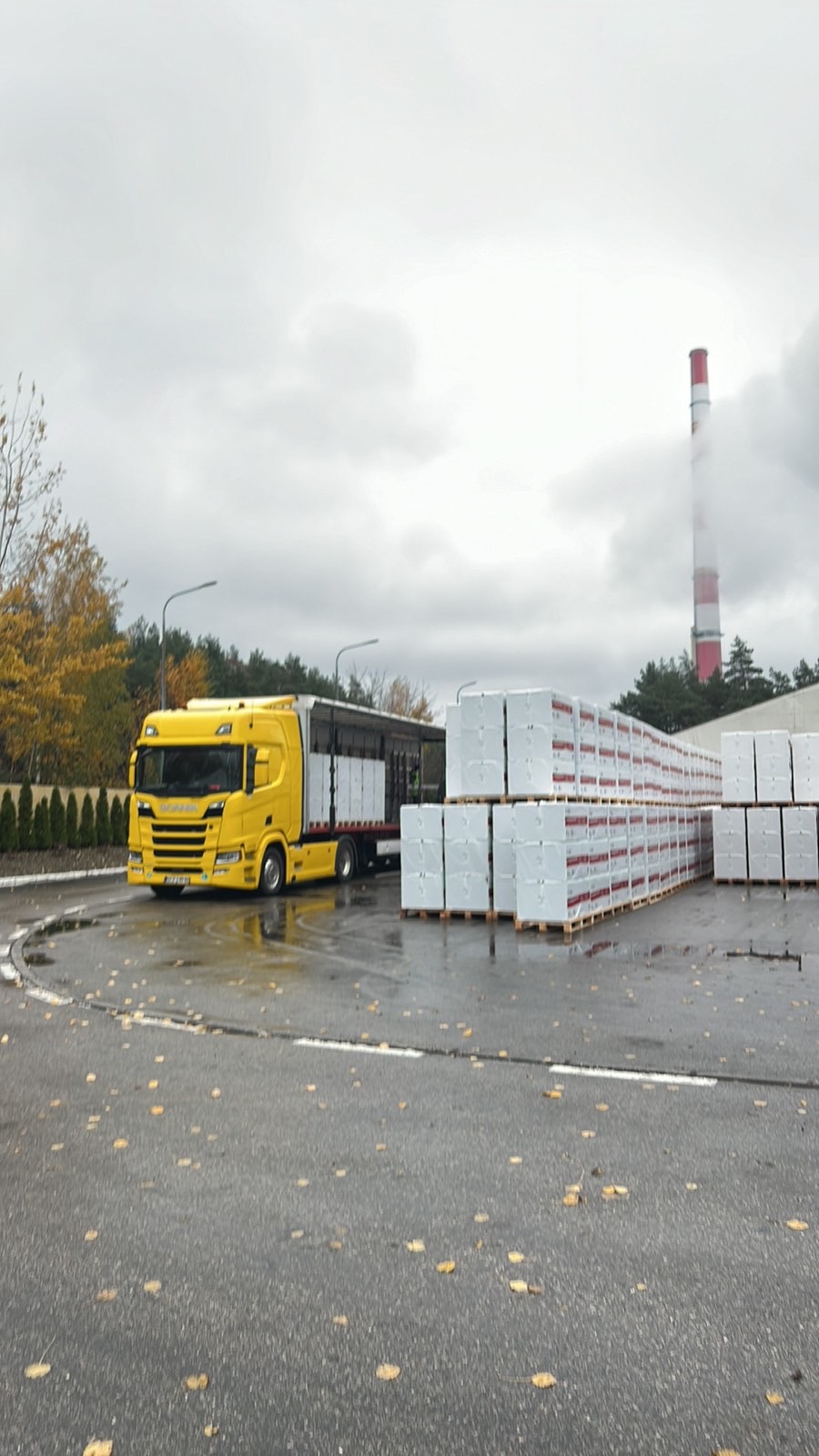 Żółta ciężarówka Scania z naczepą załadowana paletami białych bloczków budowlanych na mokrym placu, w tle komin przemysłowy i drzewa z jesiennymi liśćmi.