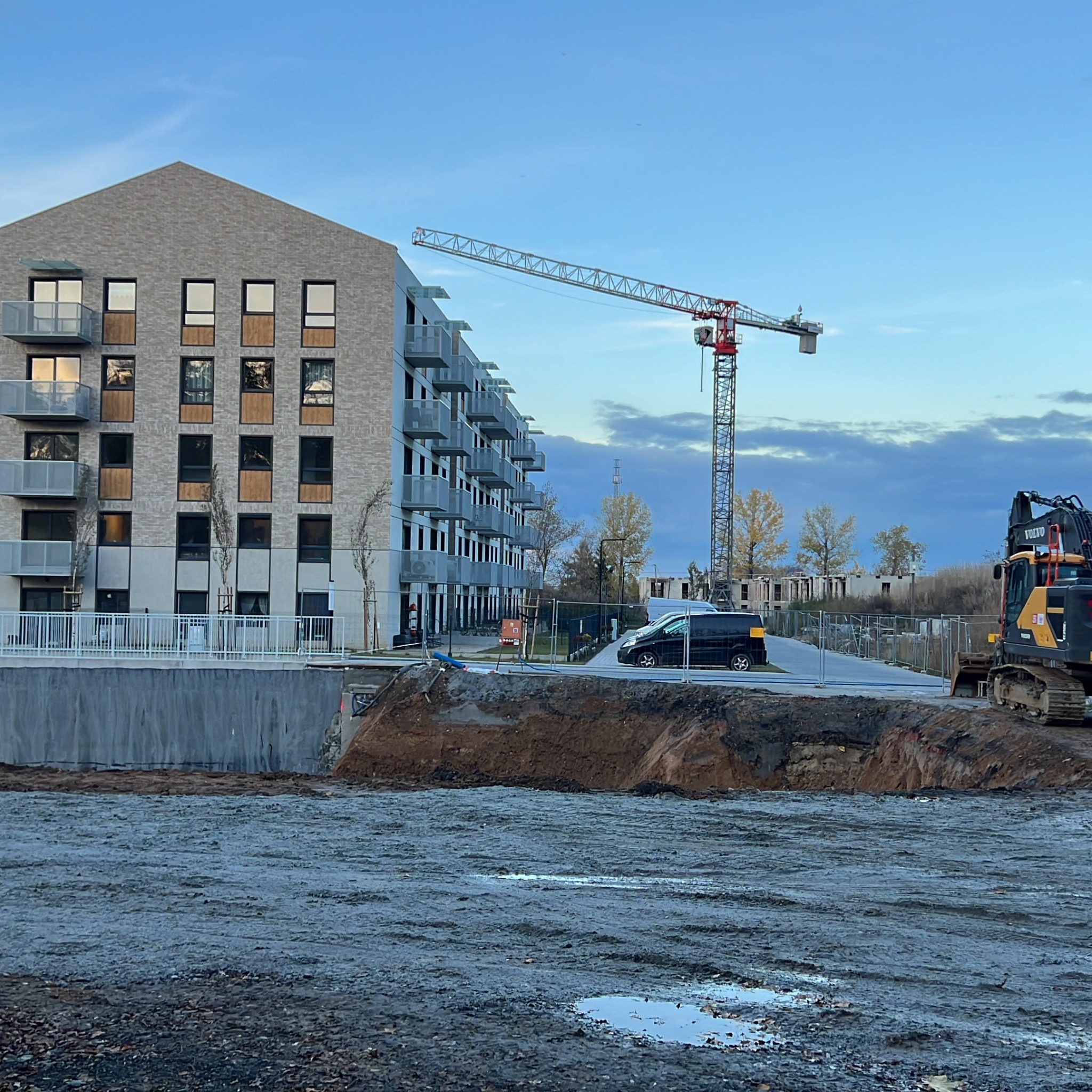 Wielopiętrowy budynek mieszkalny z jasnej cegły w trakcie budowy, widoczny żuraw budowlany i wykop z ziemią oraz ciężki sprzęt budowlany.