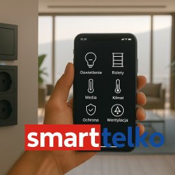 Smarttelko.pl - Smartfon z aplikacją do sterowania domem: oświetlenie, rolety, media, klimat, ochrona i wentylacja. W tle widoczne gniazdka elektryczne i okno.