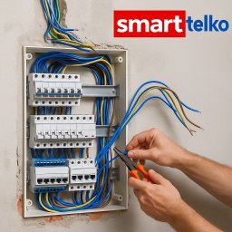 Smarttelko.pl - Elektryk przy pracy: montaż przewodów w skrzynce rozdzielczej z bezpiecznikami, używa kombinerek. Widoczne kolorowe kable i logo firmy na ścianie.