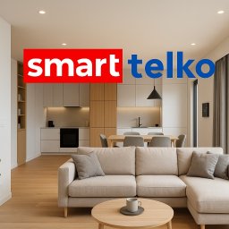 Smarttelko.pl - Nowoczesny salon z inteligentnym termostatem na ścianie i widokiem na kuchnię. Minimalistyczny design z rośliną doniczkową i przeszkleniami na ogród.