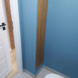 Niebieska ściana w łazience z dekoracyjnym, pionowym panelem z drewna, widoczny fragment toalety i drzwi.