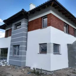 Dwukondygnacyjny dom z elewacją w kolorze białym i szarym z drewnianymi panelami na górze, okna zabezpieczone folią, widoczne pozostałości materiałów budowlanych na ziemi.