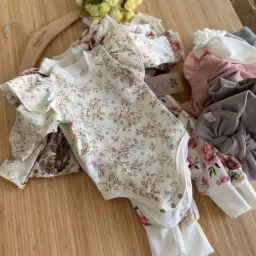 Delikatne, kwieciste body dziecięce z falbankami i dopasowane legginsy, część na wieszaku, część ułożona obok, uzupełnione o opaski w pastelowych kolorach, prezentowane przez firmę ze Szczecina.