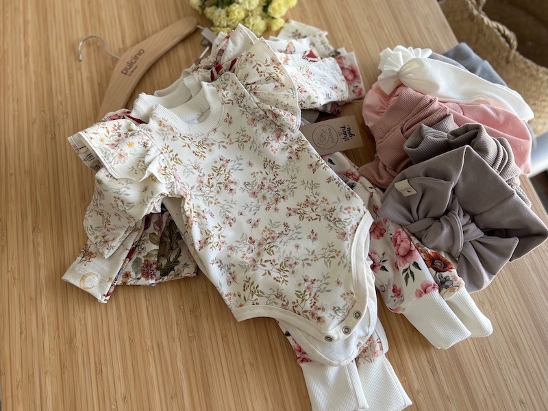 Delikatne, kwieciste body dziecięce z falbankami i dopasowane legginsy, część na wieszaku, część ułożona obok, uzupełnione o opaski w pastelowych kolorach, prezentowane przez firmę ze Szczecina.
