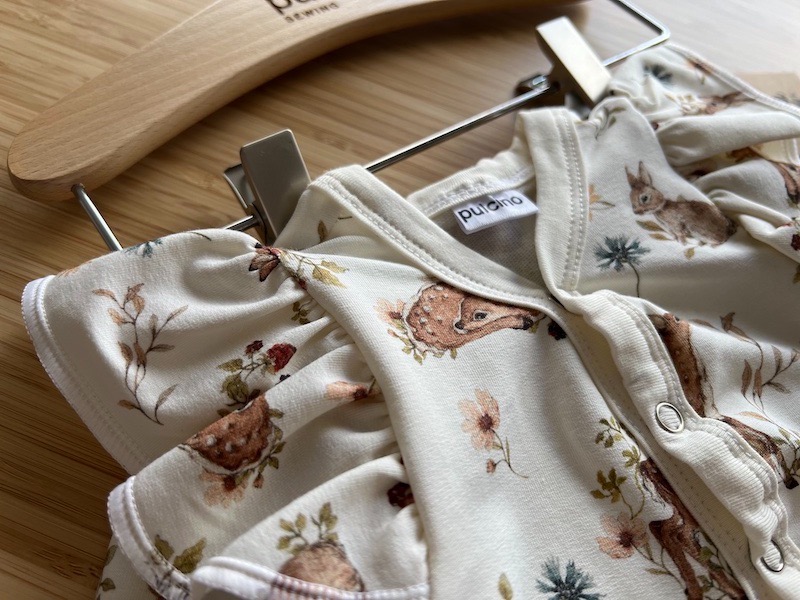 Dziecięcy pajacyk z motywem leśnych zwierząt i kwiatów, zawieszony na drewnianym wieszaku z widocznym logo marki.