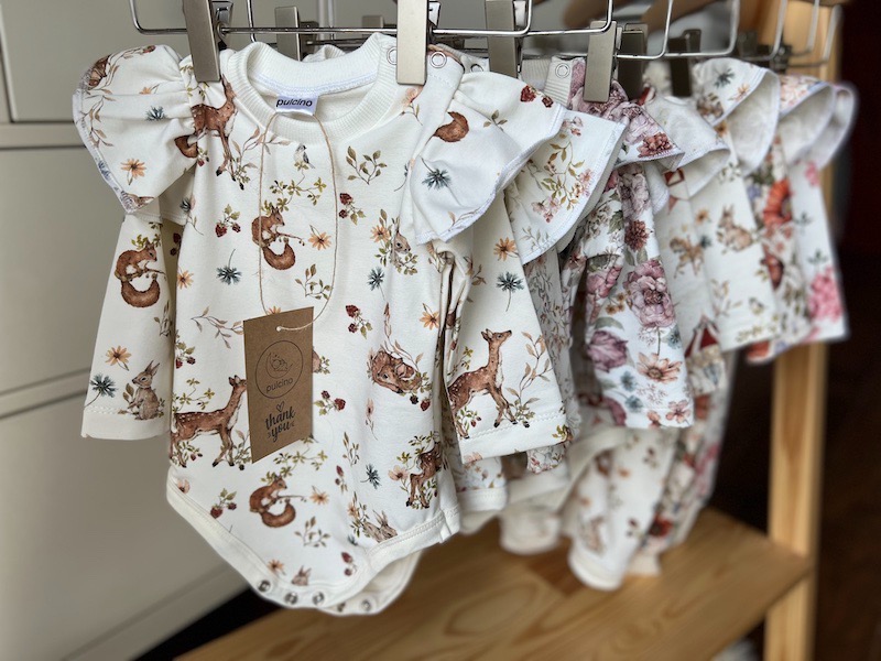 Rząd dziecięcych body z długim rękawem, z falbankami na ramionach i leśnym motywem (jelenie, wiewiórki, kwiaty), wiszących na wieszakach.