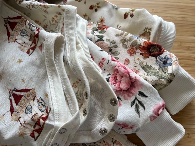 Dziecięce body z długim rękawem, z nadrukiem cyrkowym i kwiatowym, zapinane na zatrzaski, leżące na drewnianym blacie.