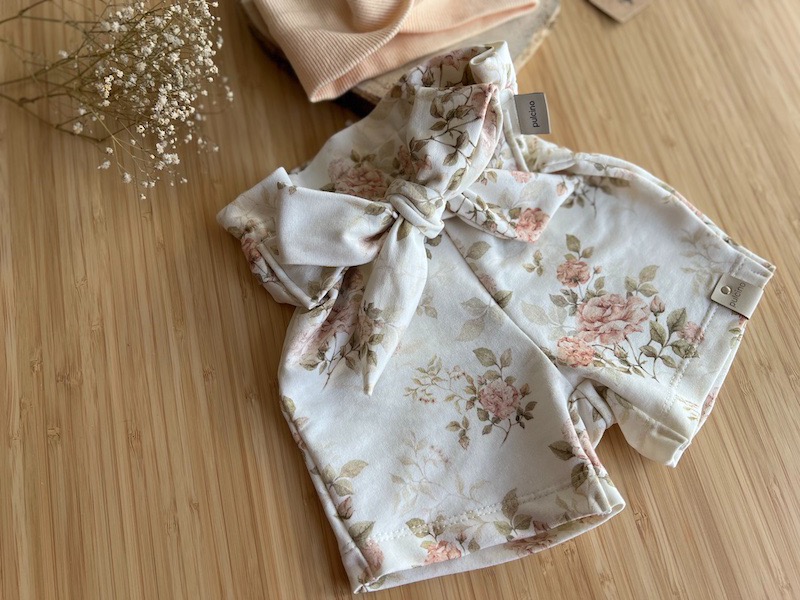 Kremowe, krótkie spodenki niemowlęce z motywem róż, przewiązane kokardą, z widoczną metką 'pulcino' na drewnianym tle.