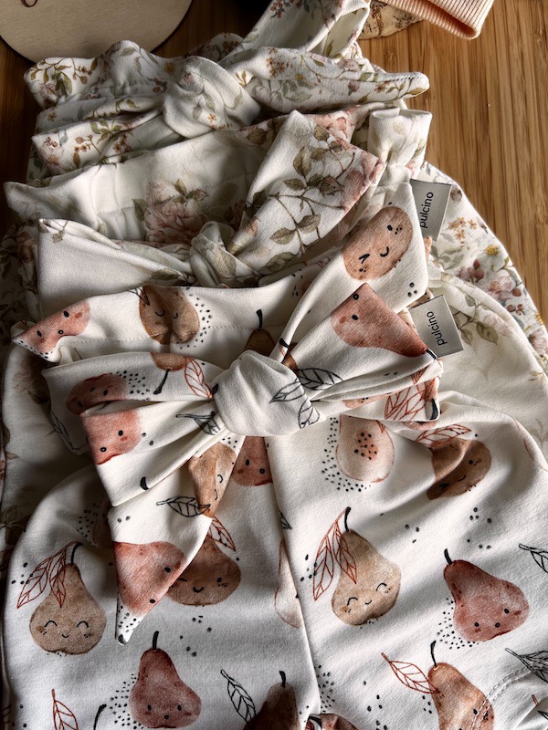 Stos ubranek dziecięcych z motywem gruszek i kwiatów, wykończonych gumką i kokardką, z widocznymi metkami.