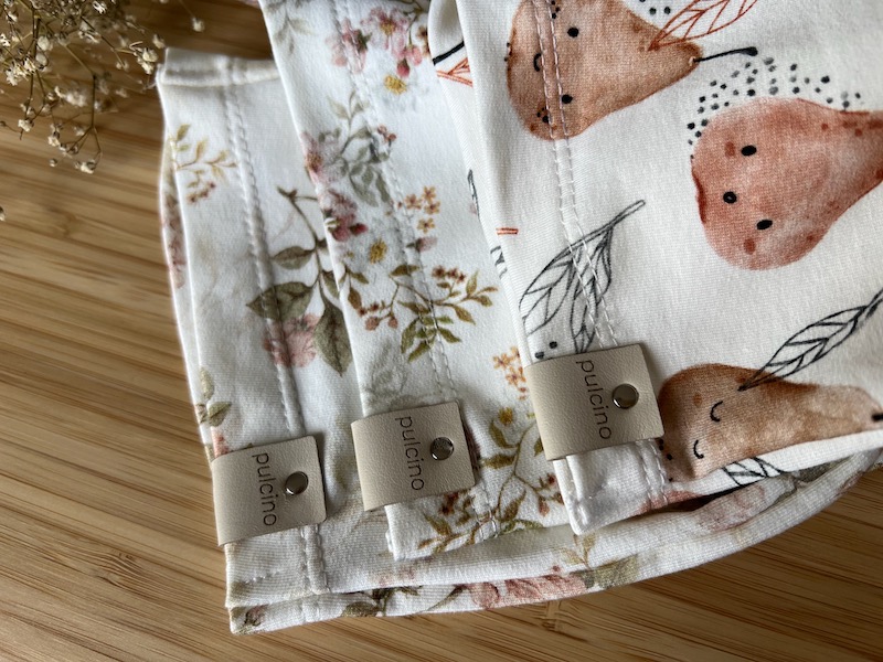 Stos trzech bawełnianych śliniaków z różnymi wzorami (kwiaty i gruszki), wykończonych szarym obszyciem i metką z napisem 'pulcino', leżących na bambusowym blacie.