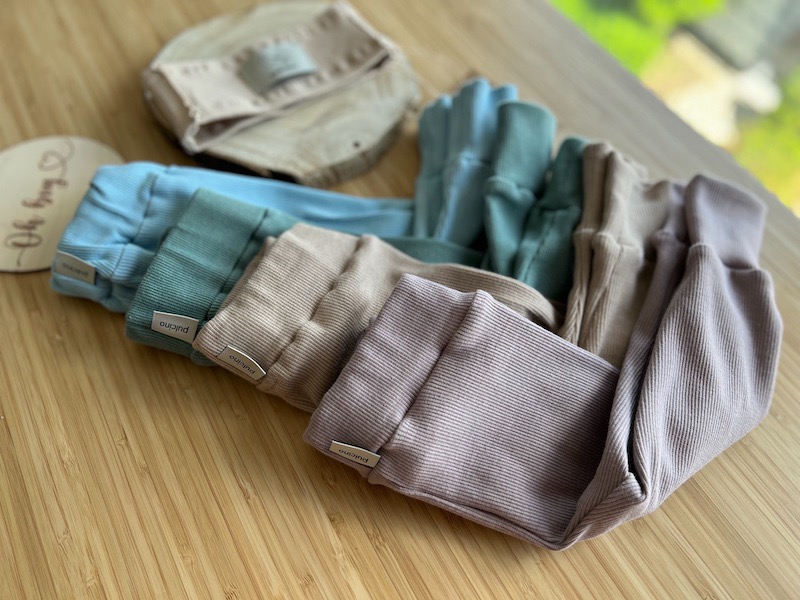 Stos pastelowych, prążkowanych spodni dziecięcych z metkami marki, ułożonych na drewnianym blacie obok opaski i dekoracyjnej drewnianej płytki.