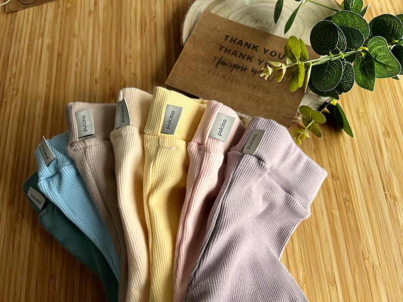 Różnokolorowe, prążkowane legginsy dziecięce z metkami marki 'pulcino', ułożone na bambusowym tle, z dekoracyjną gałązką i kartą z podziękowaniem.