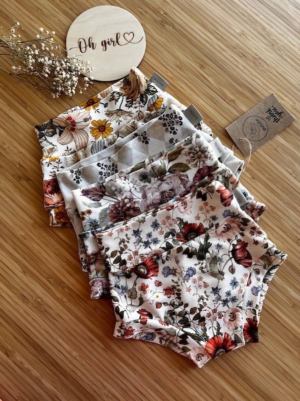 Stos pięciu par majteczek niemowlęcych z różnymi kwiatowymi wzorami ułożonych na bambusowej desce, z drewnianą zawieszką 'Oh girl' i suszonymi kwiatami w tle.