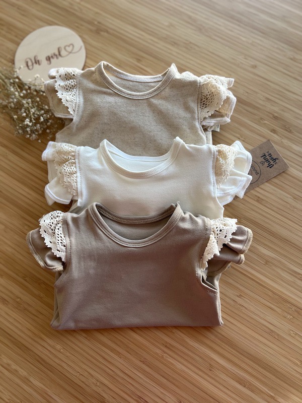 Trzy body dziecięce z koronkowymi rękawkami w odcieniach beżu, bieli i brązu, ułożone na bambusowej powierzchni, z drewnianym napisem 'Oh girl' i małą kartką z podziękowaniem.