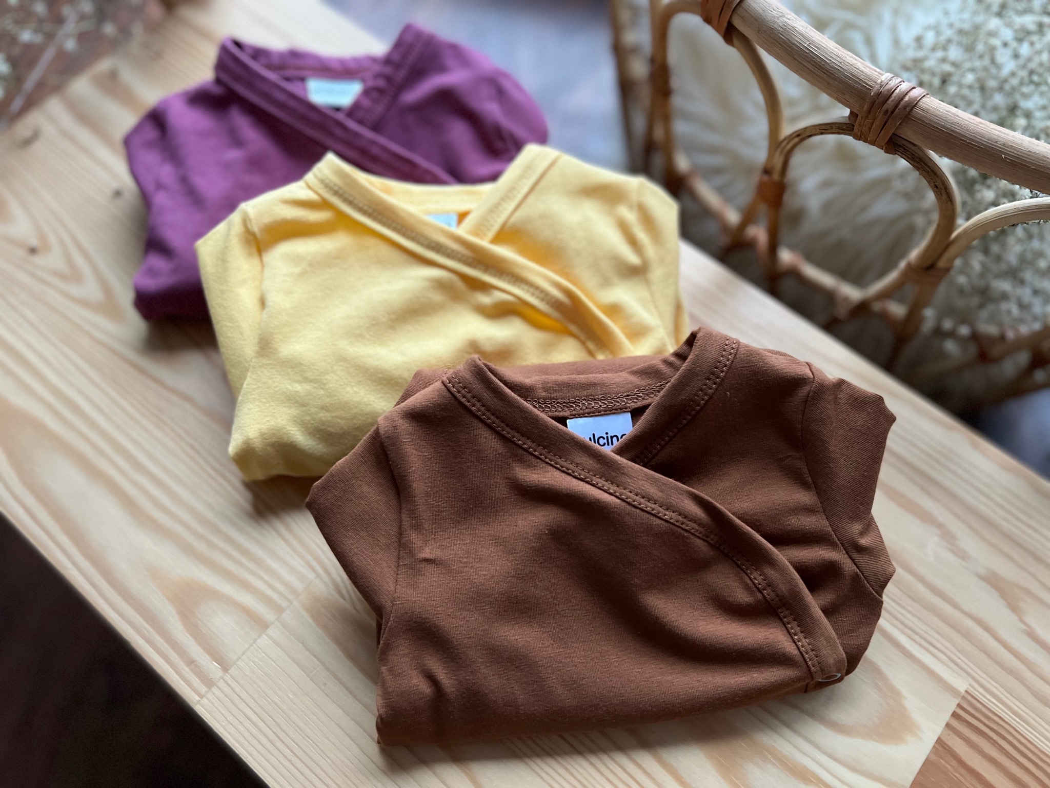 Trzy złożone body niemowlęce z krótkim rękawem w kolorach: brązowym, żółtym i fioletowym, leżące na drewnianej podłodze, z widocznym fragmentem rattanowego krzesła w tle.