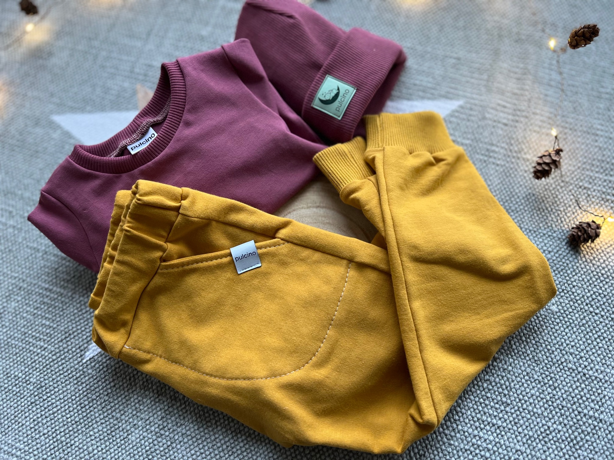 Komplet dziecięcy: bordowa bluza, musztardowe spodnie dresowe z kieszenią i bordowa czapka, leżące na szarym kocu z gwiazdą i dekoracjami z szyszek.