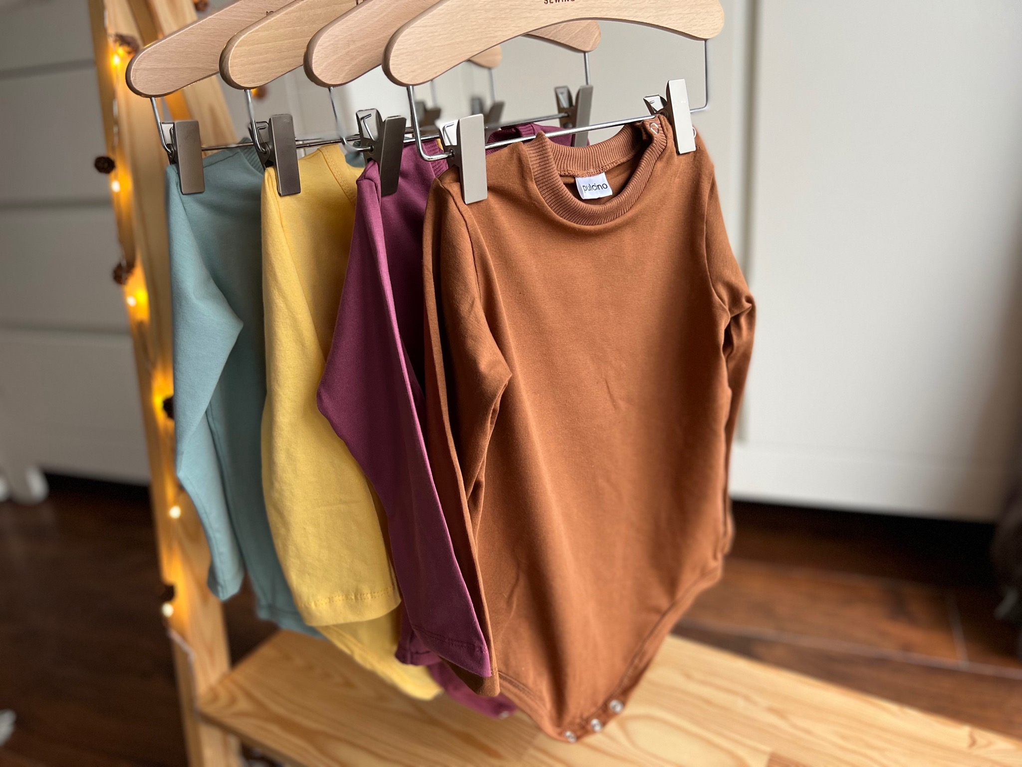 Trzy kolorowe body dziecięce (turkusowe, żółte, śliwkowe) z długim rękawem, wiszące na drewnianym stojaku z wieszakami z metalowymi klipsami, oświetlonymi delikatnymi lampkami.