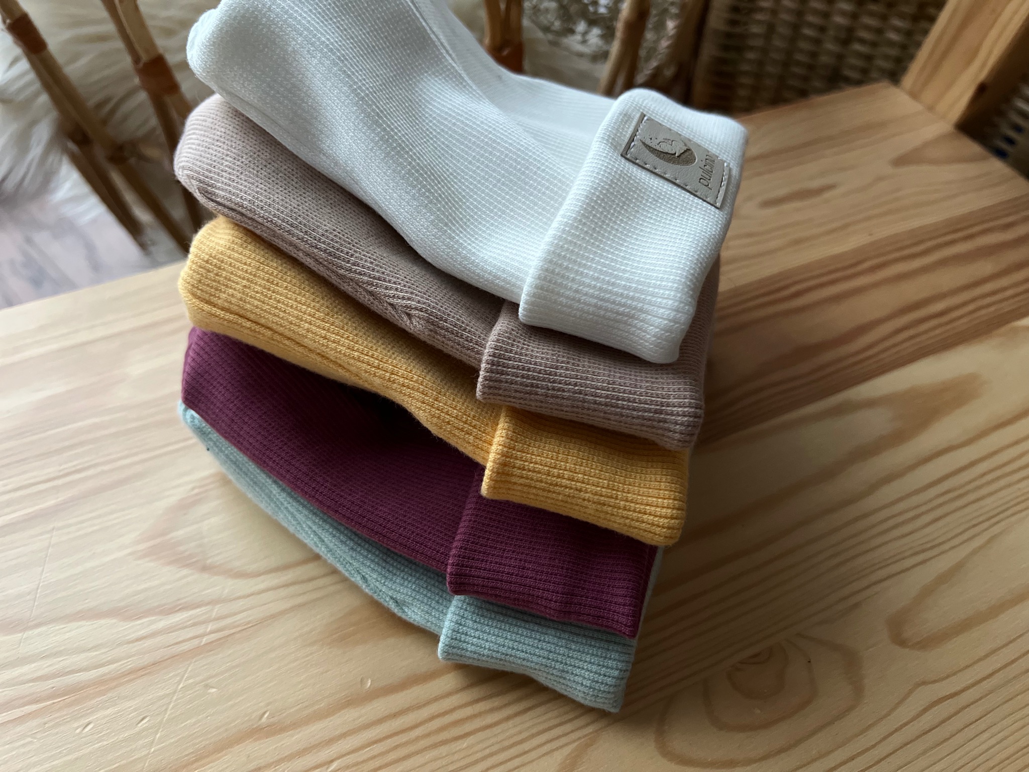 Stos pastelowych czapeczek dziecięcych z podwiniętym brzegiem i skórzaną metką na drewnianym stoliku.